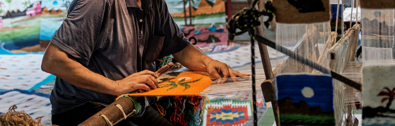Hands of middle aged man spinning colorful small rug with image of palm tree on handloom.